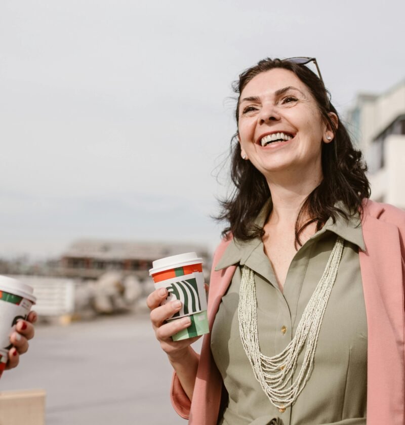 Smiling women enjoying conversation and coffee outdoors on a sunny day, promoting connection and lifestyle.