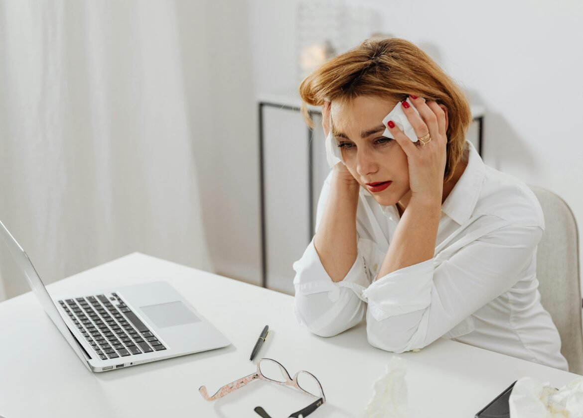 A woman in distress at her office desk, surrounded by tissues and a laptop.