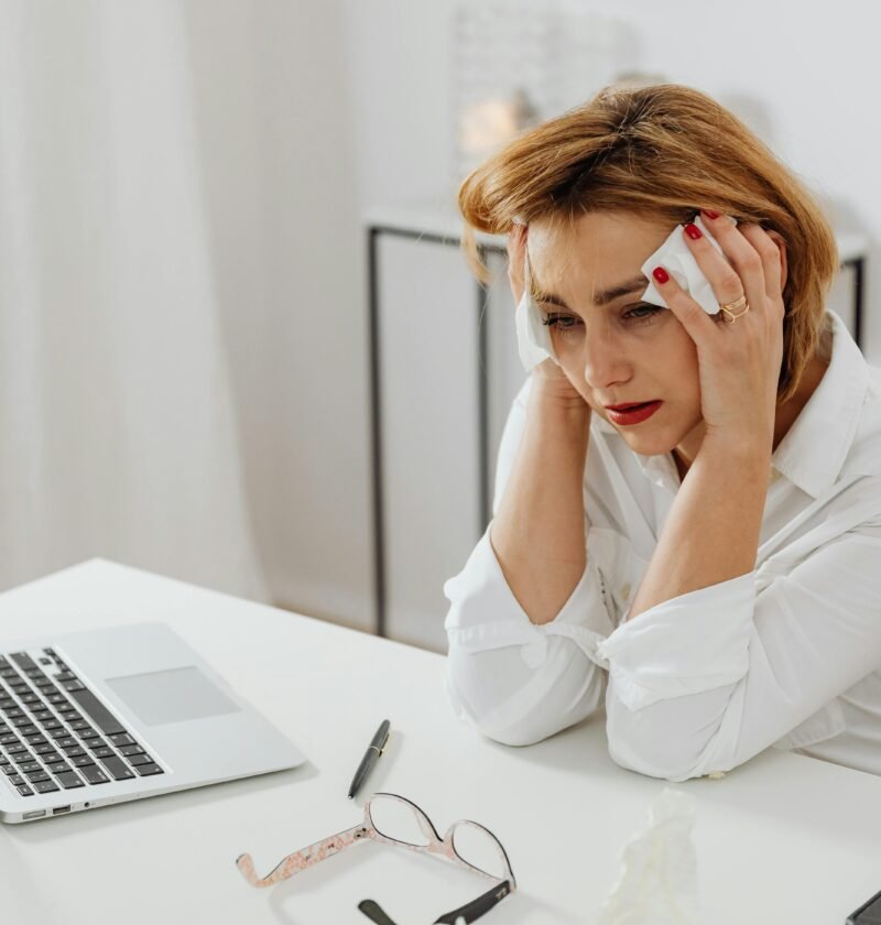 A woman in distress at her office desk, surrounded by tissues and a laptop.