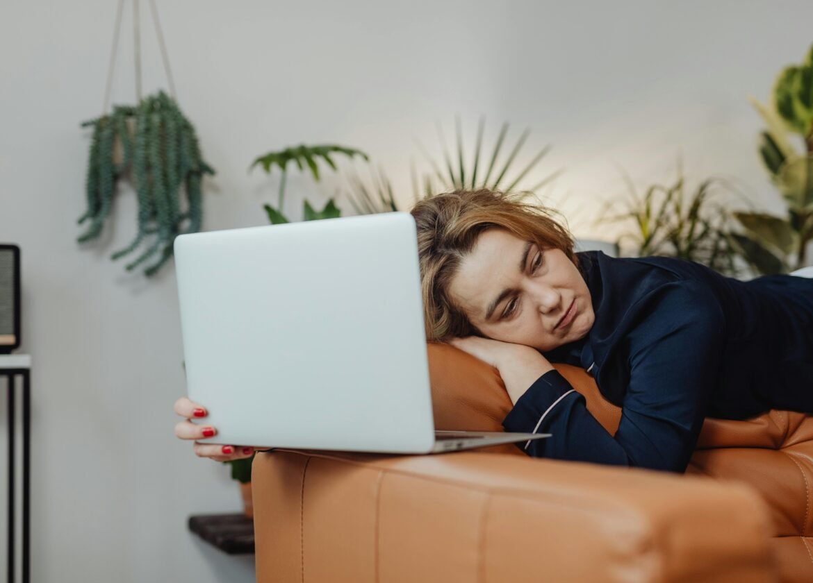 Woman sleeping on couch with laptop, surrounded by plants, indoors.
