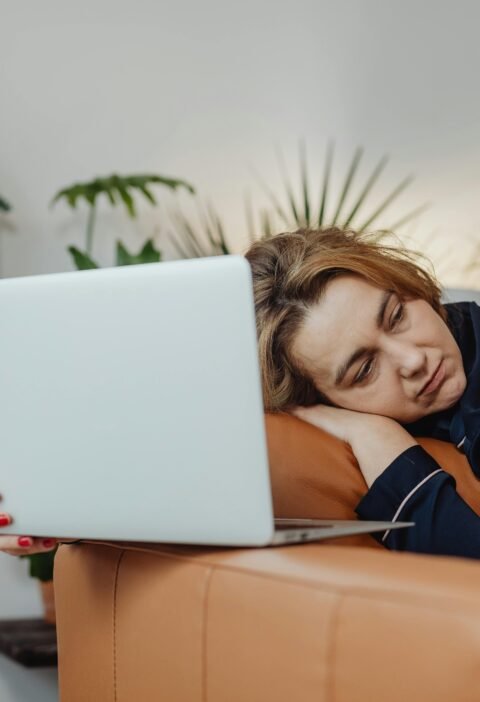 Woman sleeping on couch with laptop, surrounded by plants, indoors.