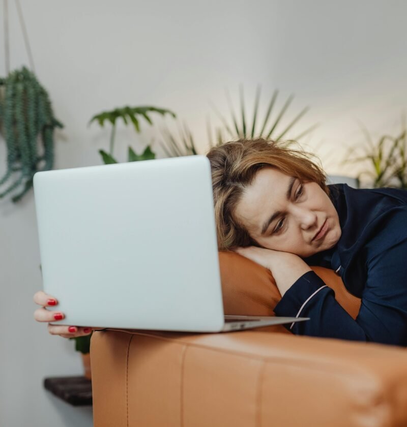 Woman sleeping on couch with laptop, surrounded by plants, indoors.
