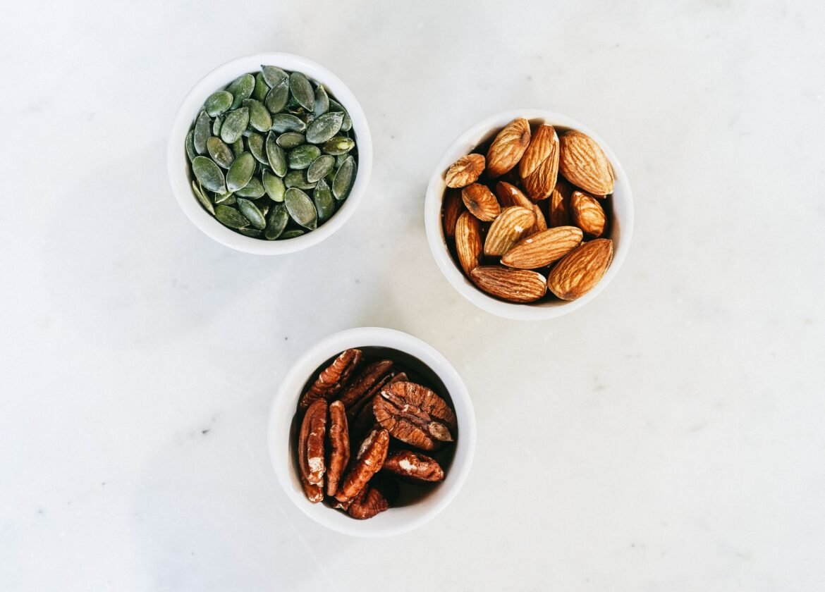A top-down view of assorted nuts in bowls, featuring almonds, pumpkin seeds, and pecans, on a marble surface.