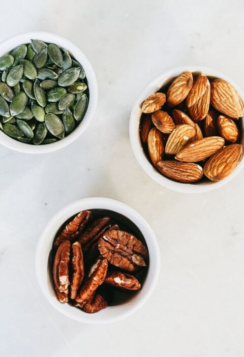 A top-down view of assorted nuts in bowls, featuring almonds, pumpkin seeds, and pecans, on a marble surface.