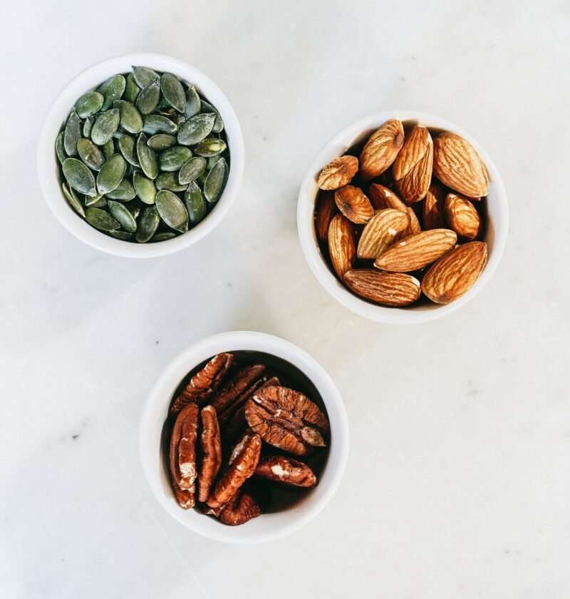 A top-down view of assorted nuts in bowls, featuring almonds, pumpkin seeds, and pecans, on a marble surface.